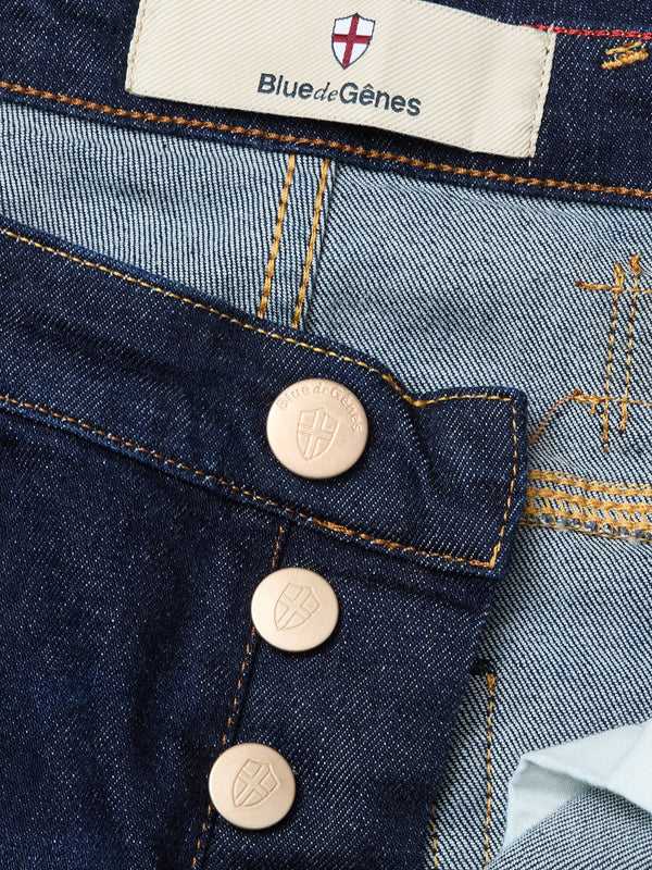 Close-up of Blue de Gênes Repi Dai V Dark V2 Jeans in dark blue denim, featuring exposed brown metal buttons, orange stitching, and a label with blue text and a red-and-white shield logo. Part of the classic five-pocket interior is visible.
