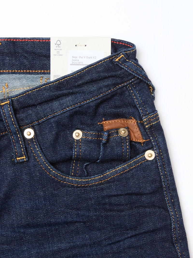 Close-up of Blue de Gênes Repi Dai V Dark V2 Jeans in dark blue denim, highlighting classic five-pocket style, copper rivets, a brown leather accent on the coin pocket, and a white tag tucked into the waistband.