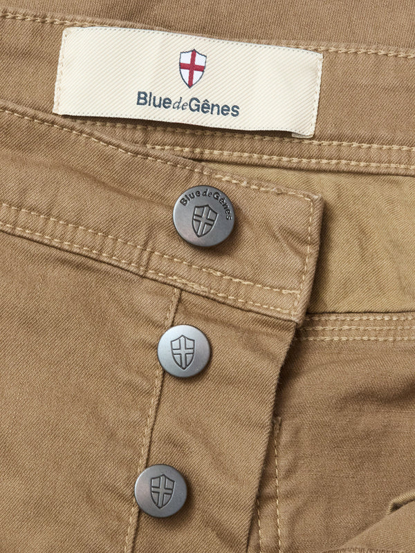 Close-up of Paulo Pavia 1 V2 Chino - Fudge by Blue de Gênes, made from cotton stretch fabric with shield-embossed metal buttons. A fabric label above the buttons displays “Blue de Gênes” and a red cross on white.
