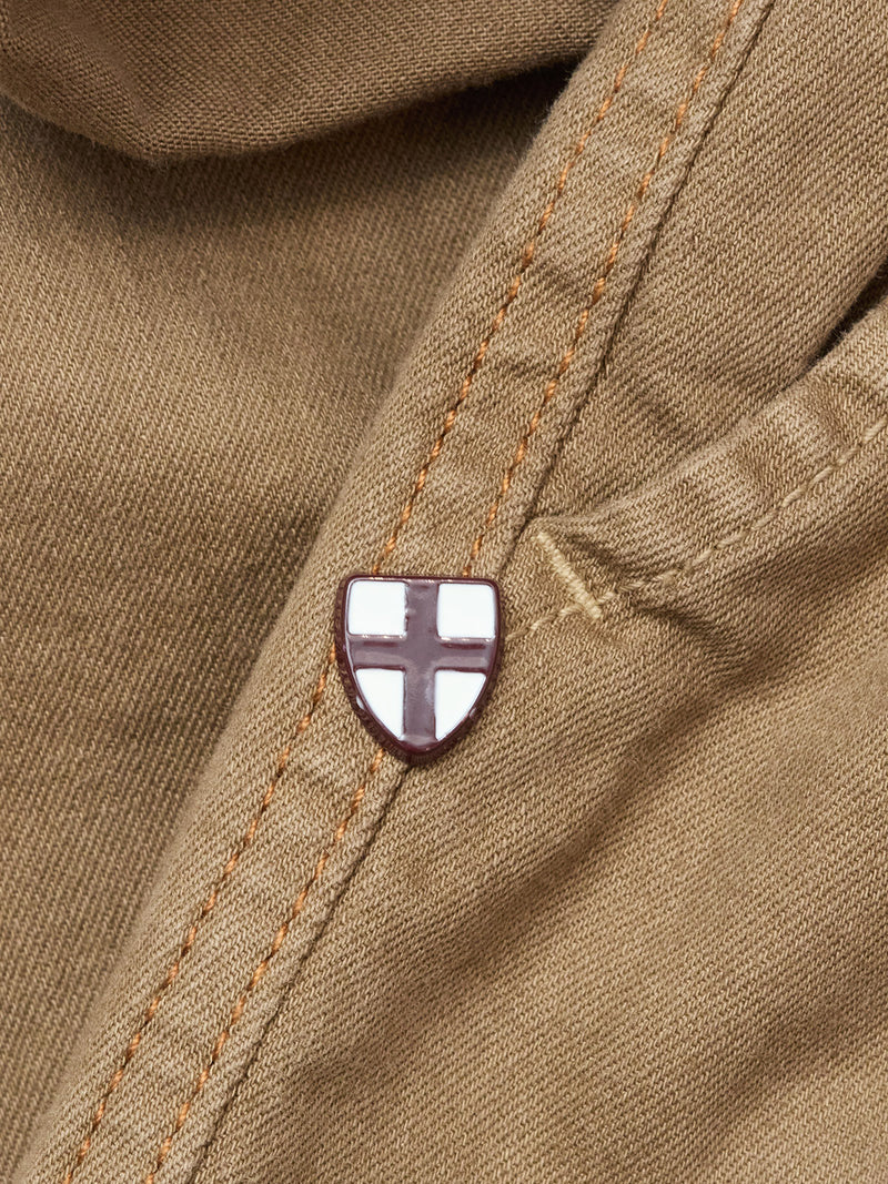 A small shield-shaped pin with a white background and red cross is attached to the seam of Blue de Gênes Paulo Pavia 1 V2 Chino - Fudge pants, adding a subtle detail to the rugged brown cotton fabric.