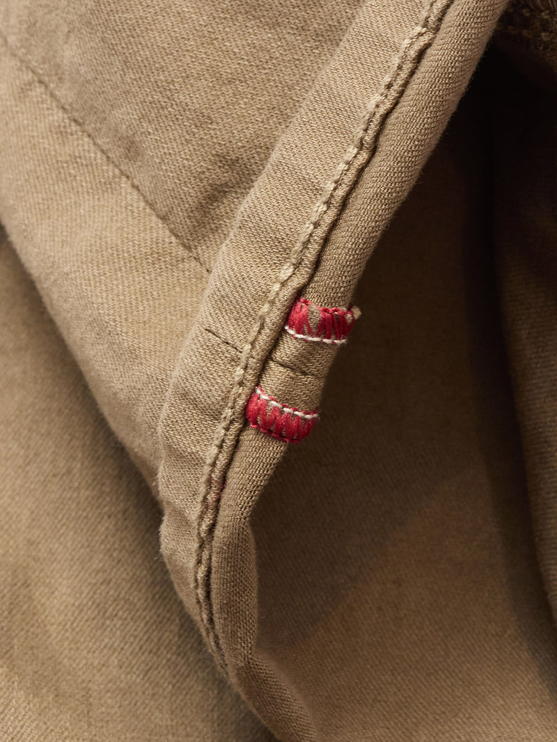 Close-up of the hem on Blue de Gênes Paulo Pavia 1 V2 Chino in Fudge, showing tan fabric with beige stitching and two small red thread sections—suggesting a repair or unique detail at the edge.