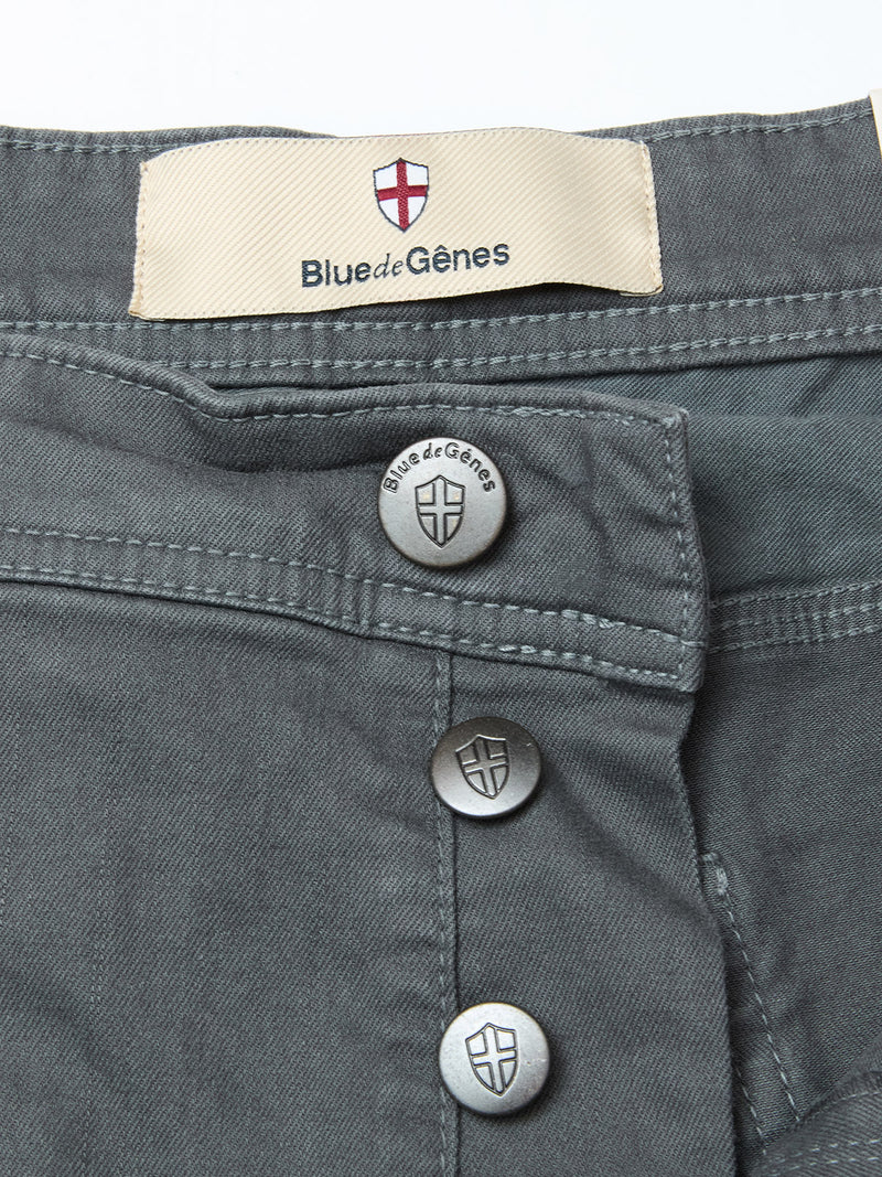 Close-up of Paulo Pavia 1 V2 Chino - Charcoal by Blue de Gênes, showing three metallic shield-logo buttons and a label with Blue de Gênes and shield emblem above the waistband. Made from cotton stretch fabric.