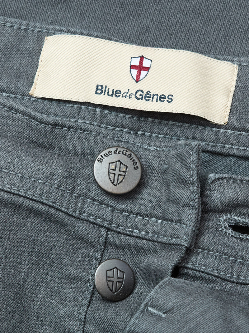 Close-up of the Paulo Pavia 1 V2 Chino in charcoal by Blue de Gênes, showcasing metal buttons and a fabric label with the brands name and shield logo, crafted from premium cotton stretch fabric.
