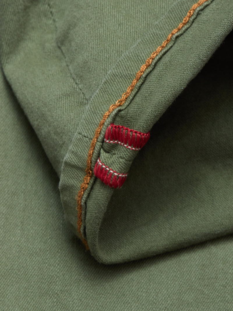 Close-up of green cotton stretch fabric with orange stitching and two small red and white stitched bars at the edge, featured on the Paulo Pavia 1 V2 Chino - Army by Blue de Gênes.
