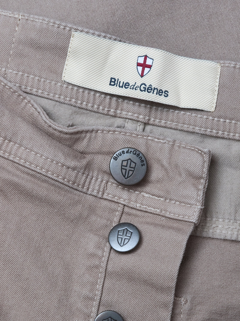 Close-up of Paulo Pavia 1 V2 Chino - Fungi by Blue de Gênes, made from light brown cotton stretch fabric. Features metal buttons and a fabric label with the Blue de Gênes name and shield logo; buttons also display the brand name and shield design.