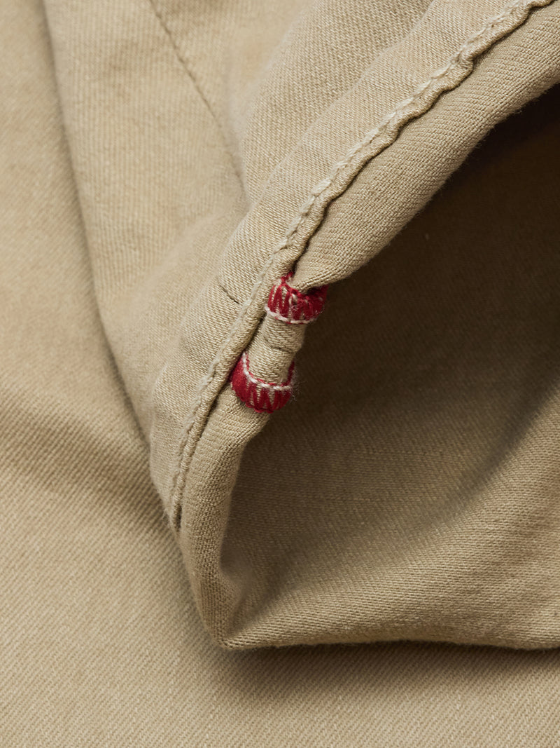 Close-up of the Paulo Pavia 1 V2 Chino in khaki by Blue de Gênes, showing beige cotton stretch fabric with visible stitching, a seam, and two small red thread accents at the hem.