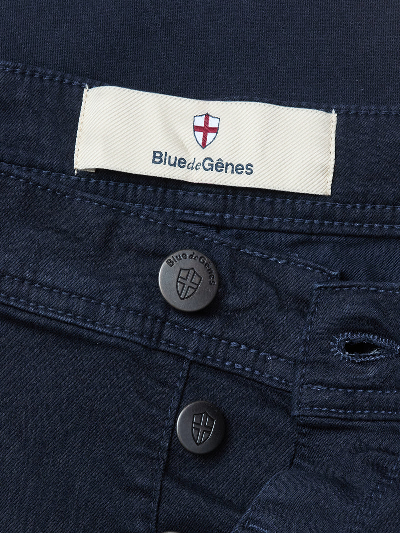 Close-up of Midnight Blue Paulo Pavia 1 V2 Chino by Blue de Gênes in cotton stretch, with a label displaying a red and white shield logo, two metal buttons featuring the shield, and subtle vintage military-inspired design details.