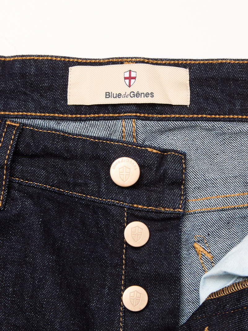 Close-up of Blue de Gênes Recco F V2 Jeans - Rinse Wash featuring dark blue denim, copper buttons, contrasting stitching, and a beige waistband label with the Blue de Gênes name and red cross shield logo.