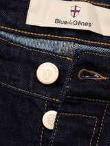 Close-up of Recco F V2 Jeans - Rinse Wash by Blue de Gênes: dark blue denim with orange stitching, two metallic buttons engraved with a shield logo, and a fabric label featuring the Blue de Gênes name and red-white shield emblem.