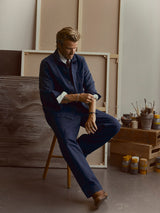 A man in a navy blue outfit sits on a wooden stool in an art studio, adjusting the buckle strap on his Blue de Gênes BGliam Nebraska Chino - Blue trousers. Art supplies and canvases are visible in the background.