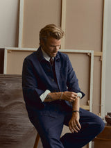 A man with light brown hair and a beard, dressed in the Blue de Gênes BGcuddy Nebraska Jacket in blue and matching pants, sits on a stool while rolling up his sleeve. Blank canvases are stacked in the background.