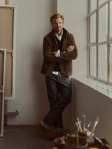 A man with blond hair and a beard stands by a window in an art studio, dressed in a brown jacket, white shirt, and Blue de Gênes BGleroy Jeans - Raw Denim. He stands with arms crossed; paintbrushes sit in the foreground.