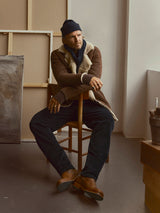 A man in a brown shearling jacket, navy beanie, and Blue de Gênes BGpalco Cord Chino - Navy sits on a wooden chair in an art studio, surrounded by canvases and art supplies.
