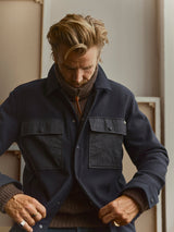 A man with blond hair looks down while buttoning the Blue de Gênes BGBoyd Wool Jacket in Dark Navy over a brown turtleneck sweater. He stands indoors with window light and artwork or boards in the background.