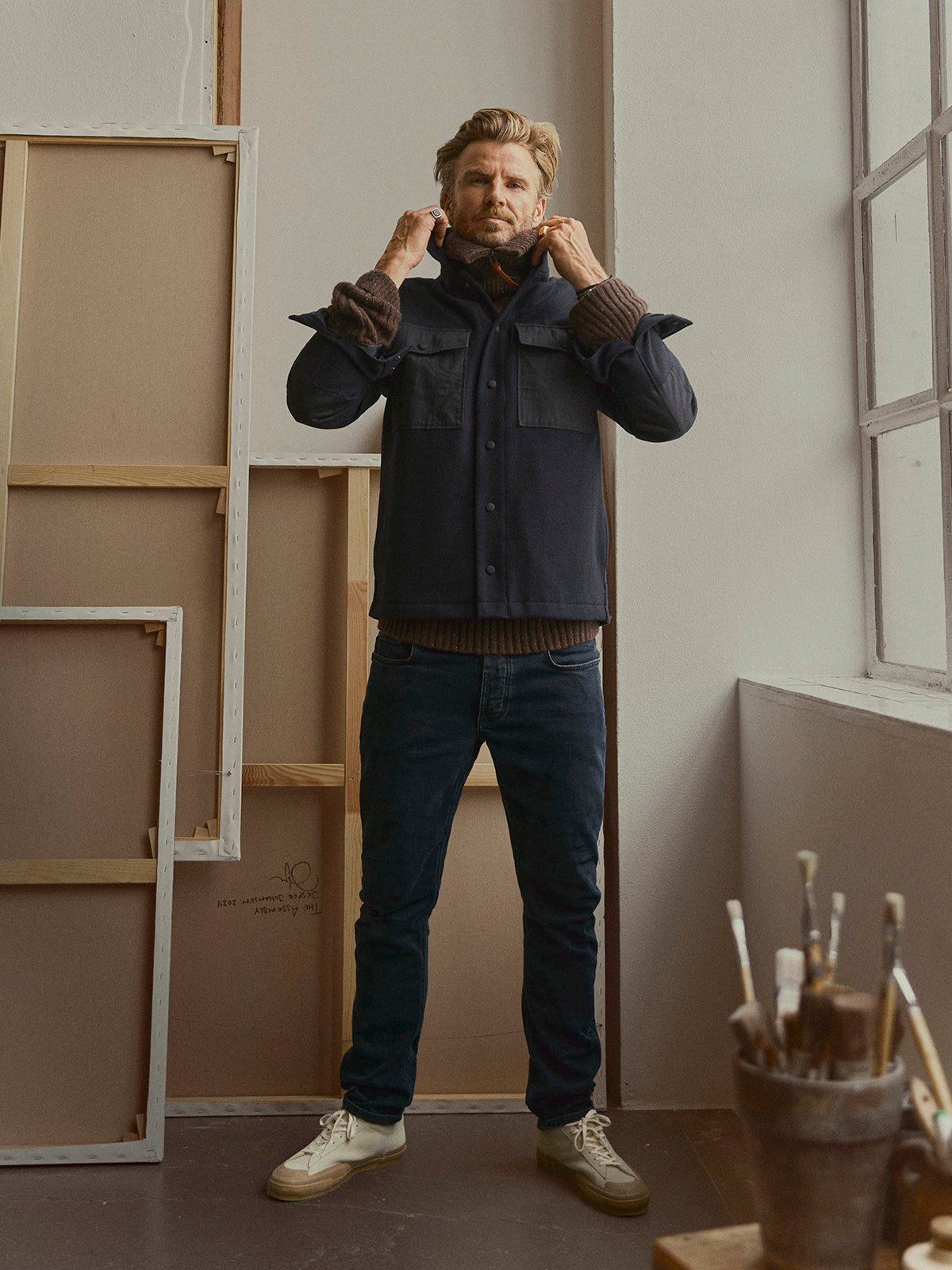 A man stands indoors by empty picture frames, wearing a dark jacket over a brown sweater, BGvinci Black OD Jeans in Blue Black Denim from Blue de Gênes, and white sneakers. Paintbrushes in a jar sit on a nearby windowsill.