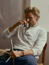 A man with blond hair sits on a chair, sipping from a mug and holding paintbrushes in one hand. He wears the BGgreg L/S Grandad shirt in white by Blue de Gênes, paired with blue pants, bracelets, and a scarf. A painting canvas appears in the background.