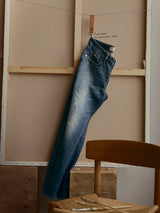 A pair of BGvinci Break Jeans - Denim wash by Blue de Gênes, featuring slight distressing and crafted from comfort stretch denim, hangs from a wooden clothespin on a canvas stretcher frame, with a wooden chair and box in the foreground.