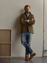 A man with folded arms leans against a wall, wearing a brown blazer, layered shirts, and Blue de Gênes BGvinci Break Jeans in a classic denim wash, paired with brown boots. An empty picture frame rests on the floor nearby.