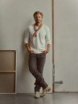 A man with a beard stands against a white wall, wearing a white shirt, Blue de Gênes BGvinci Iro Pant in Hot Fudge Brown (regular fit), tan boots, and an orange scarf. His hands are in his pockets, appearing relaxed in a minimal, studio-like setting that highlights the comfort stretch twill of his pants.