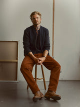 A man with blond hair and tattoos sits on a wooden stool, wearing a dark jacket, patterned scarf, Blue de Gênes BGpalco Cord Chino trousers in Ginger Bread Orange, and brown shoes in a minimal studio space.