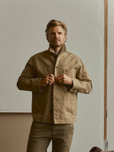 A man with light hair and a mustache stands indoors, wearing the Blue de Gênes BGcuddy Nebraska Jacket in brown—crafted from high-density cotton with a subtle dobby weave. He buttons his collar while looking slightly away from the camera, his matching pants completing the utilitarian ensemble.
