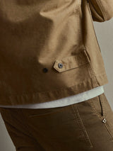 A close-up shot showcases a person wearing the BGcuddy Nebraska Jacket in brown by Blue de Gênes, paired with matching brown pants. The jacket is crafted from high-density cotton with a subtle dobby weave, and a white shirt peeks out underneath. The image highlights the distinct clothing textures and intricate pocket details.