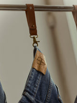 A pair of BGrepi Presley Jeans - Denim Blue by Blue de Gênes hangs from a leather strap on a metal rod. These regular slim-fit jeans feature a brown patch adorned with a stitched geometric logo. The softly blurred background keeps the focus on the jeans and strap.