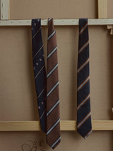 Three neckties hang on a wooden frame: the BGshield Tie - Dark Navy by Blue de Gênes, featuring navy stripes with small crest patterns; one solid brown silk tie accented with thin stripes; and a dark-hued option decorated with orange and tan diagonal stripes.