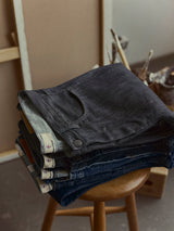 A stack of folded Blue de Gênes BGleroy Jeans - Raw Denim in various shades sits on a wooden stool, with art supplies and canvases blurred in the background.