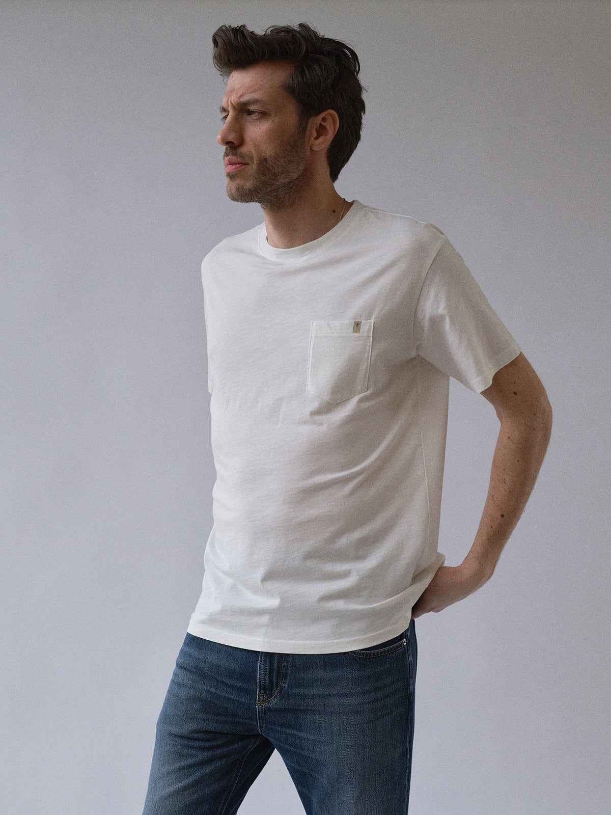 A man with short brown hair and light facial hair stands against a plain background, wearing the Blue de Gênes BGoscar T-Shirt - Snow Fall, a 100% cotton crewneck with a pocket, and blue jeans, looking thoughtfully to his left.