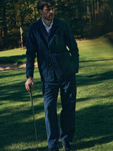 A man wearing the Blue de Gênes BGkendrick Toll Jacket in Navy Stripe stands on a golf course holding a club, with trees and a flag visible in the background.