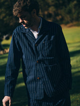 A man wears the Blue de Gênes BGkendrick Toll Jacket in Navy Stripe, standing on grass outdoors as sunlight casts shadows on him, with blurred trees in the background.