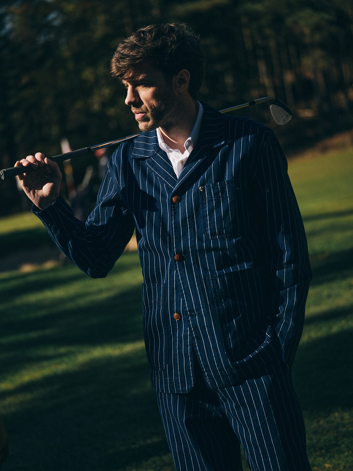 A man in a Blue de Gênes BGkendrick Toll Jacket - Navy Stripe holds a golf club over his shoulder while standing on a sunny golf course with trees in the background.