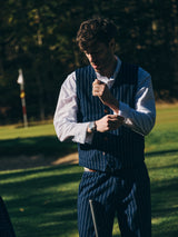 A man in a pinstripe suit adjusts his Blue de Gênes BGsander Oxford Shirt - White while standing on grassy terrain with trees and a distant flag, suggesting a possible golf course setting.