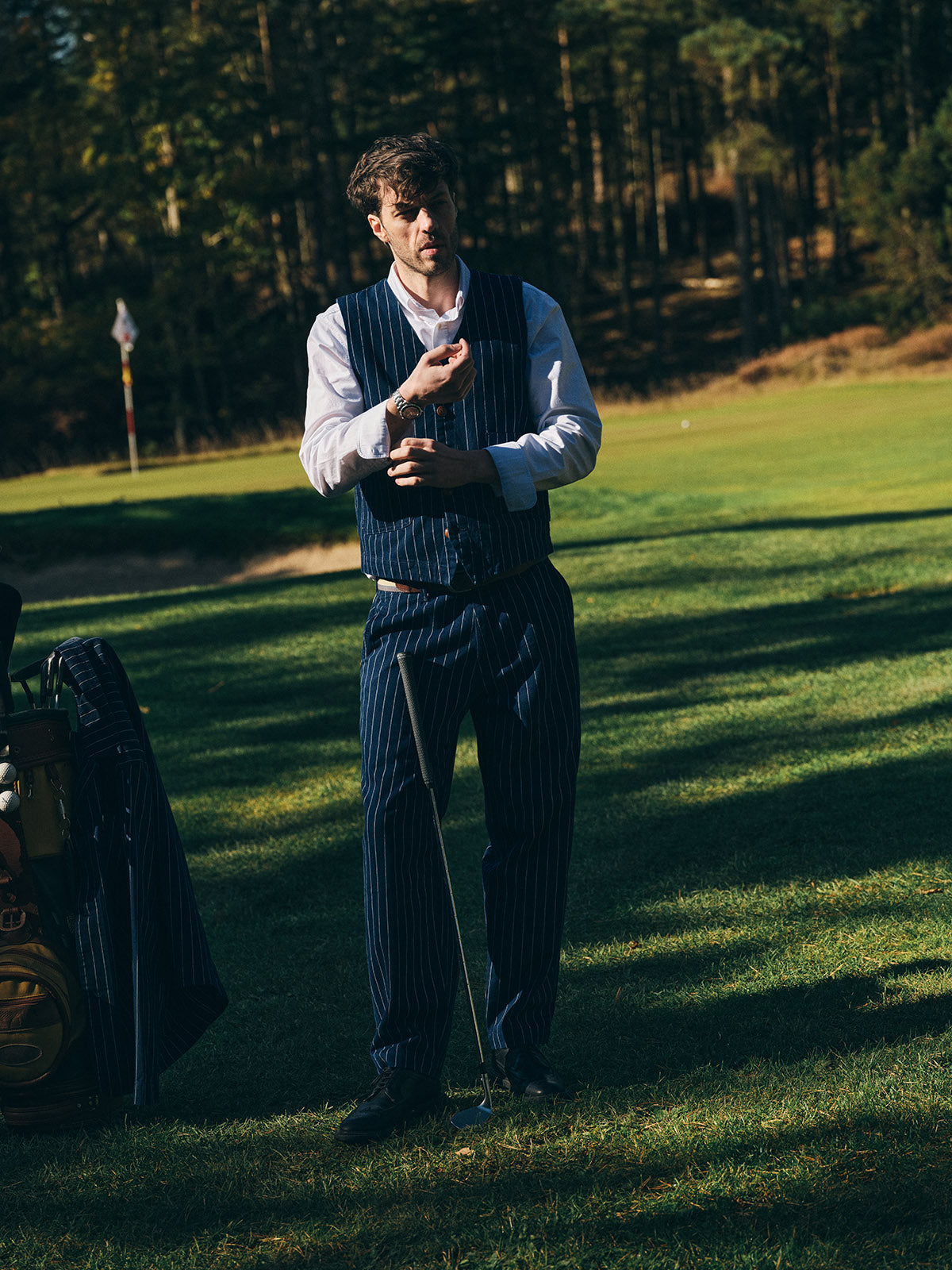 A man in a pinstripe vest and Blue de Gênes BGpalco Toll Pants - Navy Stripe stands on a golf course, adjusting his watch as the sun casts shadows over the grass, with trees and a golf flag visible in the background.