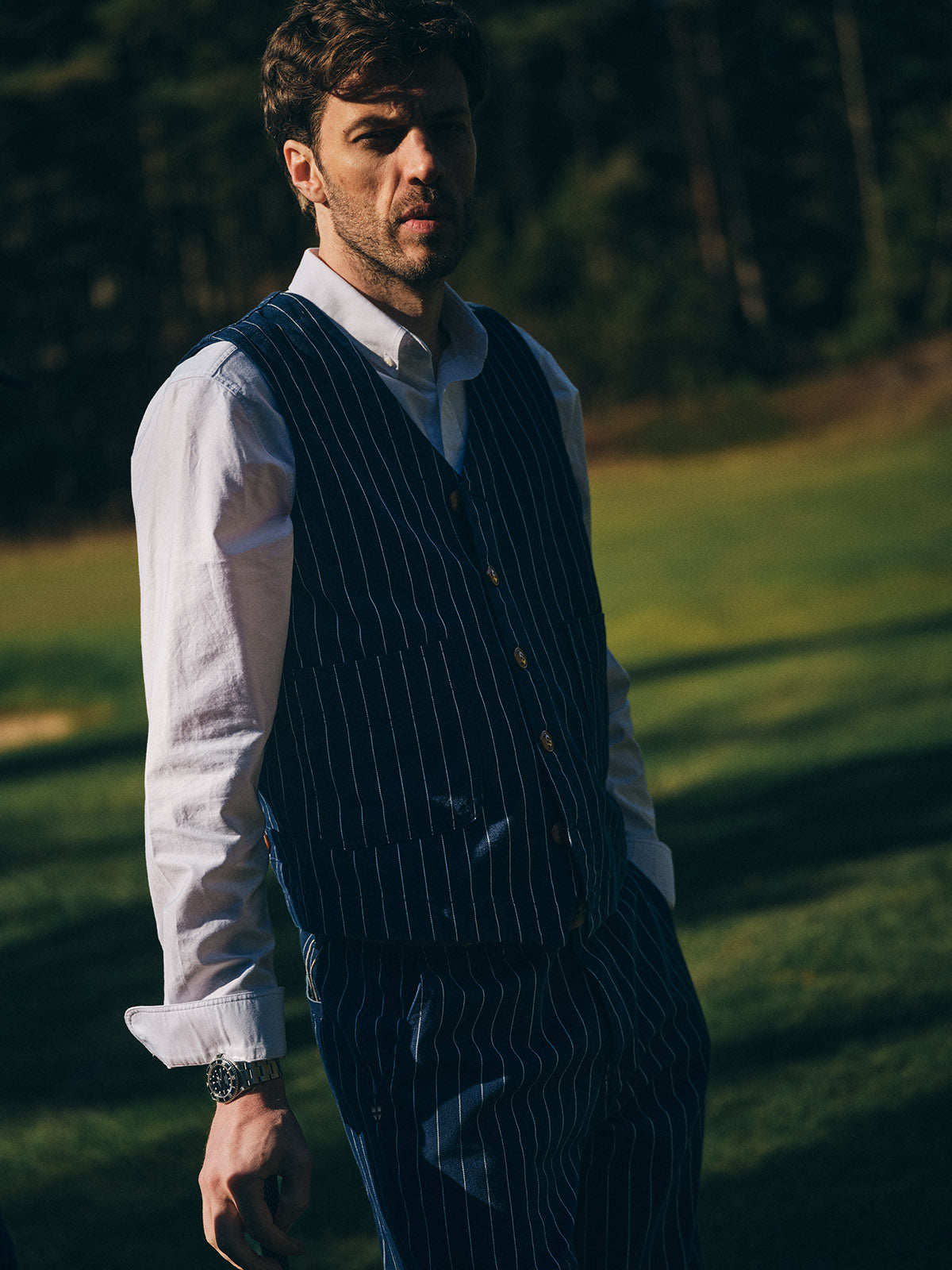 A man stands on a grassy field under the sun, wearing the Blue de Gênes BGsander Oxford Shirt in white, paired with a blue pinstripe vest and trousers; trees form the background as sunlight casts shadows on his face.