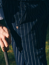 A close-up of a person wearing Blue de Gênes BGpalco Toll Pants - Navy Stripe while holding a golf club, with sunlight casting shadows and a small metallic badge visible on the pants.