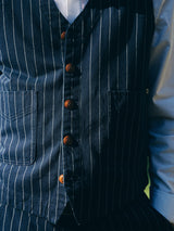 Close-up of a person wearing the Blue de Gênes BGlumen Toll Gilet - Navy Stripe, a worker-inspired, striped cotton vest with brown buttons and two front pockets, layered over a light blue dress shirt. The face is not visible.