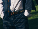 Close-up of a person in a blue pinstripe suit, white shirt, tan belt, and BGpalco Toll Pants - Navy Stripe by Blue de Gênes. Hands in pockets, showcasing elevated outdoor workwear style on grass.