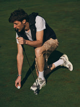 A man in casual golf attire kneels on a green, holding a golf club and placing a ball. He wears BGcupra Print Shorts - Multi Pattern by Blue de Gênes, a white t-shirt, vest, white socks, and sneakers.