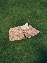 The BGcupra Print Shorts - Multi Pattern by Blue de Gênes are laid flat on a grassy lawn under sunlight, showing off their pink and beige cotton twill with white lining.