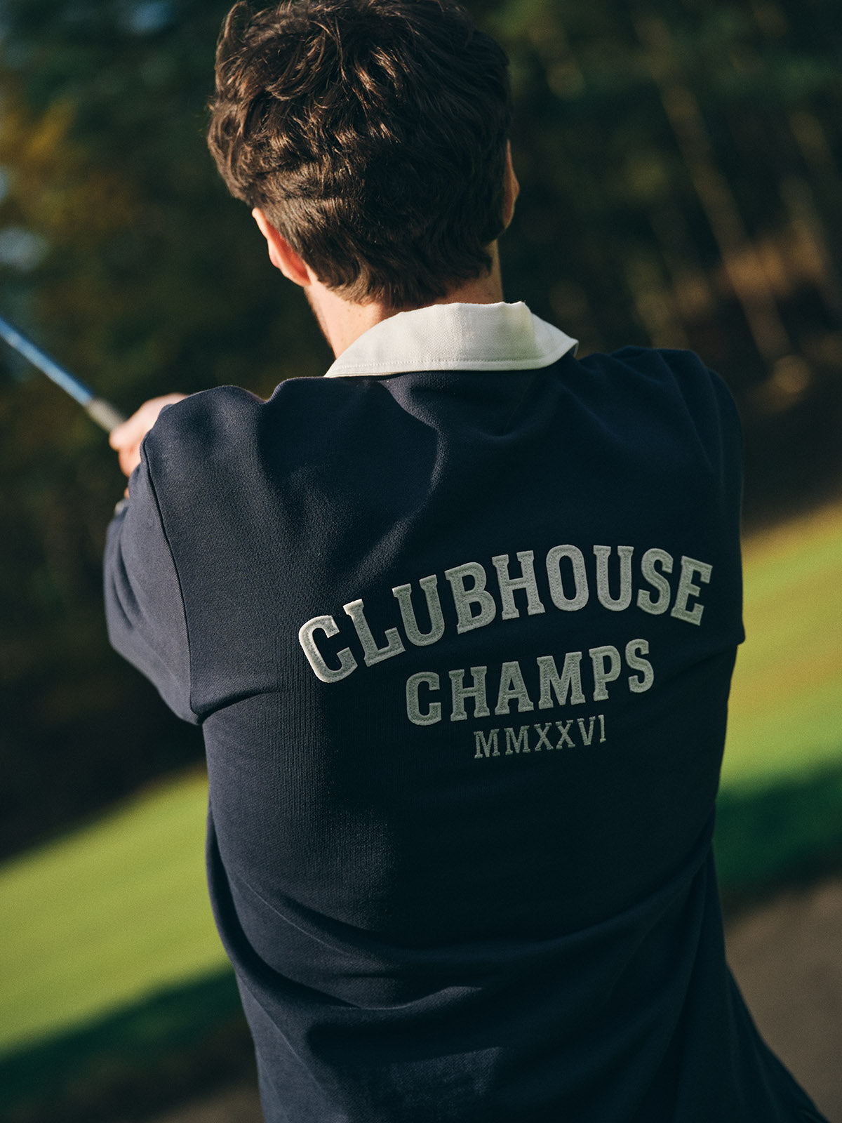 A person seen from behind, outdoors with blurred trees, holds a golf club while wearing a Blue de Gênes BGclubhouse Rugby Sweatshirt - Navy from the Clubhouse Champs Collection, featuring 