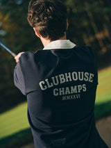 A person seen from behind, outdoors with blurred trees, holds a golf club while wearing a Blue de Gênes BGclubhouse Rugby Sweatshirt - Navy from the Clubhouse Champs Collection, featuring "CLUBHOUSE CHAMPS MMXXVI" printed on the back.