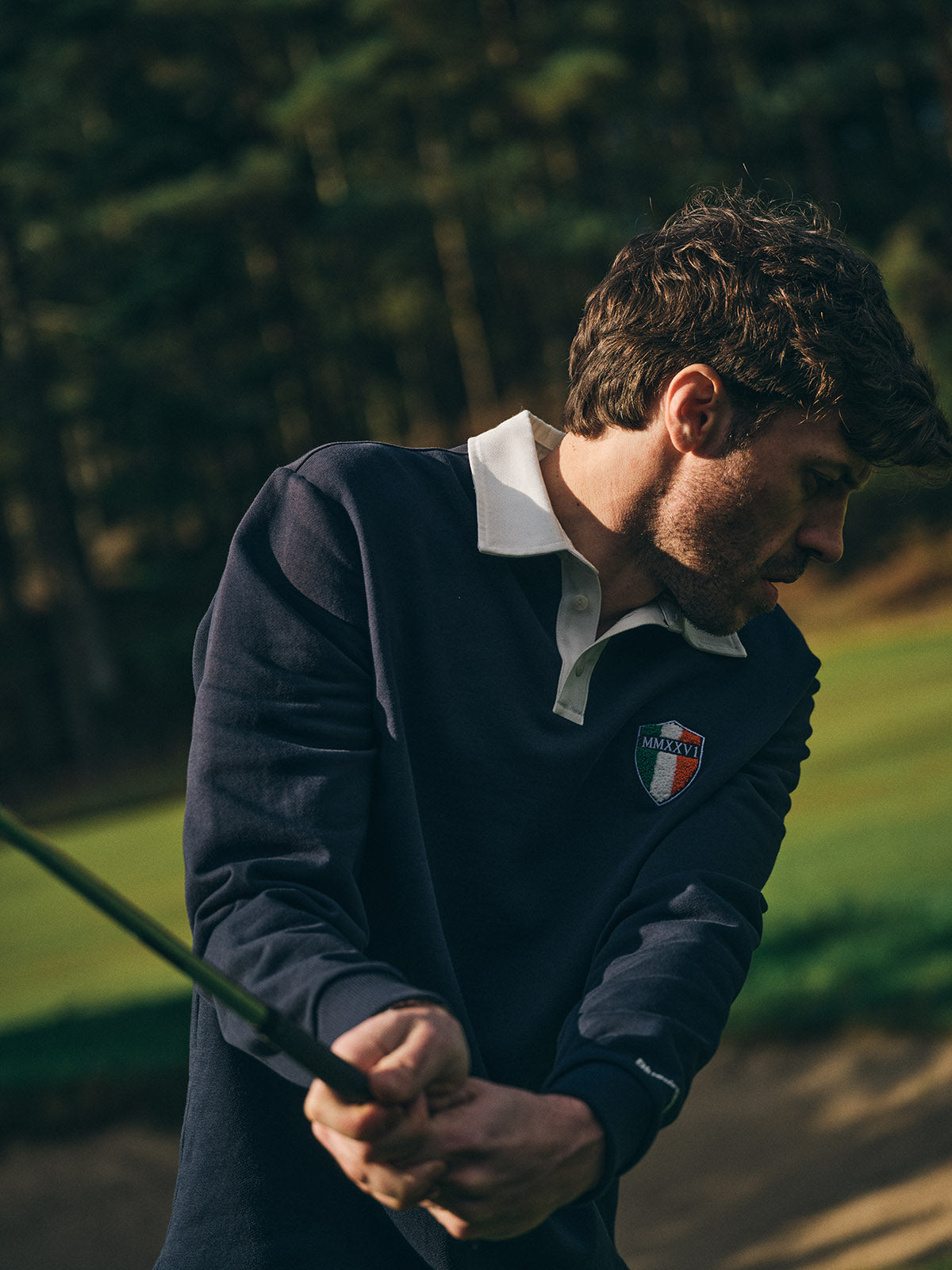 A man wears the Blue de Gênes BGclubhouse Rugby Sweatshirt in navy as he swings a golf club on a course, with blurred trees and grass in the background.