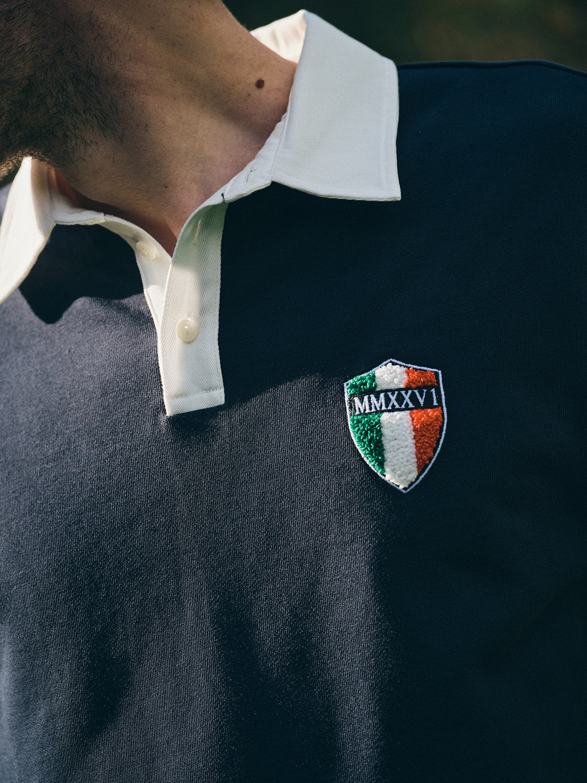 A person wears the BGclubhouse Rugby Sweatshirt - Navy by Blue de Gênes, featuring a white collar, buttons, a crest patch with Italian flag colors, and MMXXVI embroidery on the chest. Part of the Clubhouse Champs Collection.