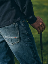 A person in a dark sweatshirt and Blue de Gênes BGvinci Striker Jeans - Dark Distress holds a golf club on green grass, with the image highlighting their hand, jeans pocket, and the club handle.