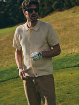 A man with facial hair and sunglasses, wearing a beige short-sleeve knit shirt, BGgiacomo Color Jeans - Tanin by Blue de Gênes, and a golf glove stands on a grassy golf course with trees, holding something in his mouth.