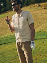 A man in sunglasses, a cream knit shirt, tan pants, and a golf glove smiles on the golf course as he tosses a ball—his BGgiacomo Color Jeans - Tanin from Blue de Gênes add a casual touch to his sporty look.