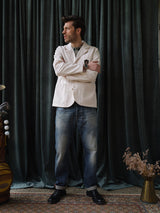 A man stands before dark green curtains, wearing a white blazer, light gray shirt, and Blue de Gênes BGgiacomo Tim Jeans in Antique wash. He looks to the side with arms crossed; a rug, brass vase, and dried flowers lie beside him.