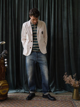 A man stands indoors on a patterned rug before dark green curtains, wearing a white blazer, striped shirt, black shoes, and Blue de Gênes BGgiacomo Tim Jeans - Antique wash. A golf bag and dried flowers lie beside him.